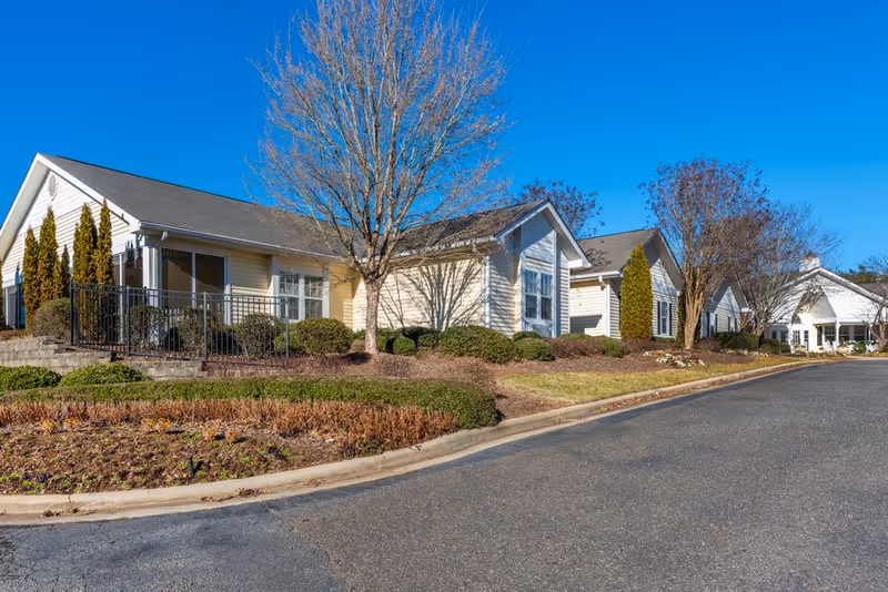 Single-story beige residential buildings with landscaped shrubs and leafless trees lining a curved driveway under a clear blue sky.