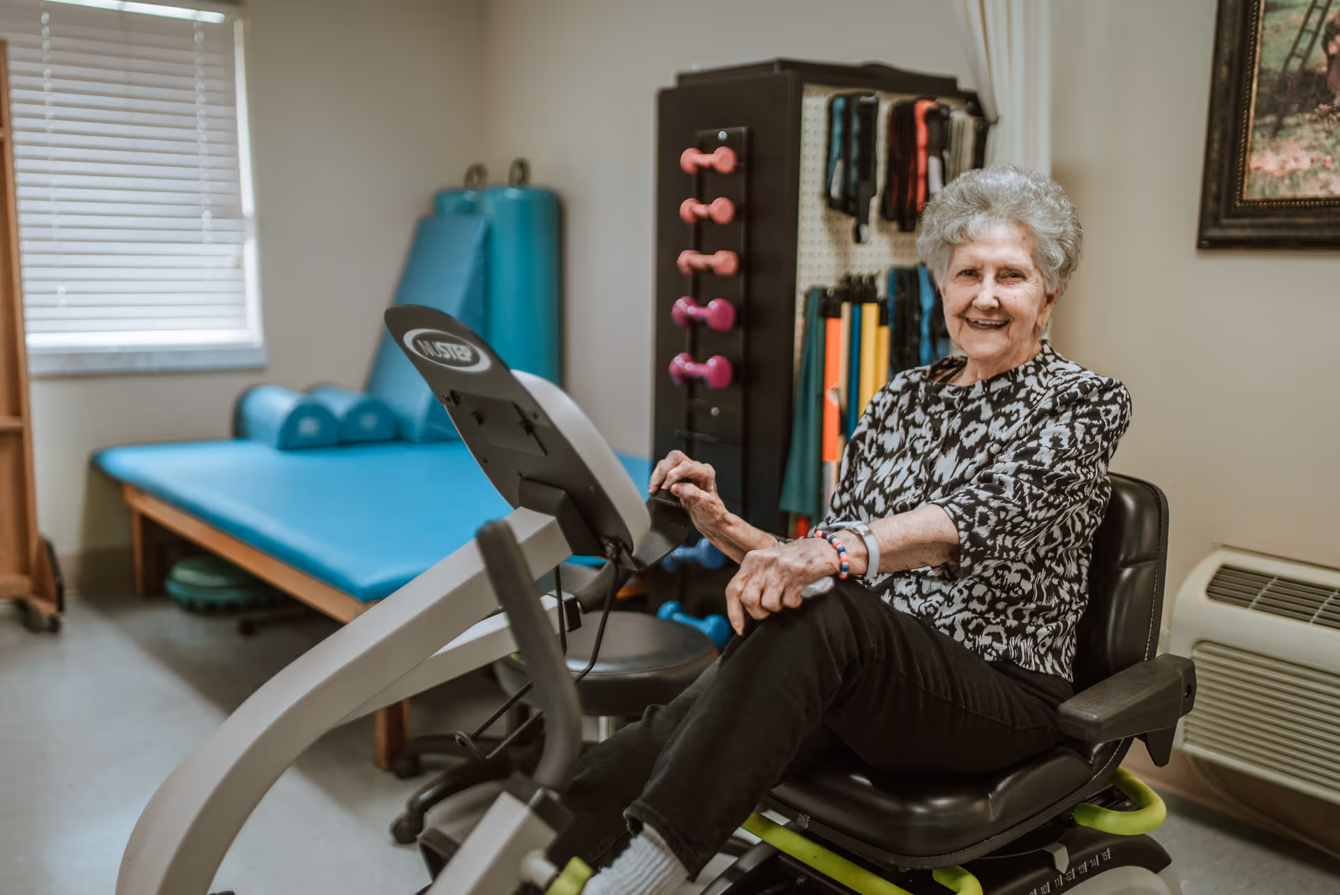 An elderly woman sitting on a recumbent exercise bike in a room with exercise equipment including dumbbells, resistance bands, and a padded therapy table. She is smiling and appears to be in a fitness or rehabilitation area.