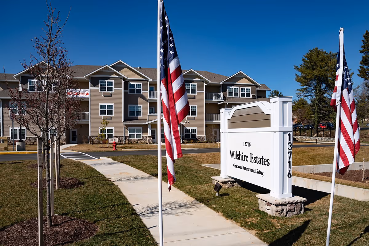 Exterior view of Wilshire Estates Gracious Retirement Living community building with a clear blue sky, a sidewalk leading to the entrance, two American flags on poles, young trees planted along the sidewalk, and a large white sign displaying the community name and address 13716.