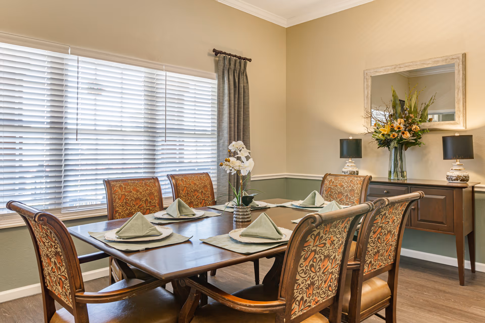 A dining room with a wooden table set for six people. Each place setting has a green napkin folded on a white plate with a green placemat. The chairs have patterned upholstery with floral designs. A window with white blinds and a gray curtain is on the left wall. On the right wall, there is a wooden sideboard with two lamps and a large floral arrangement beneath a rectangular mirror.