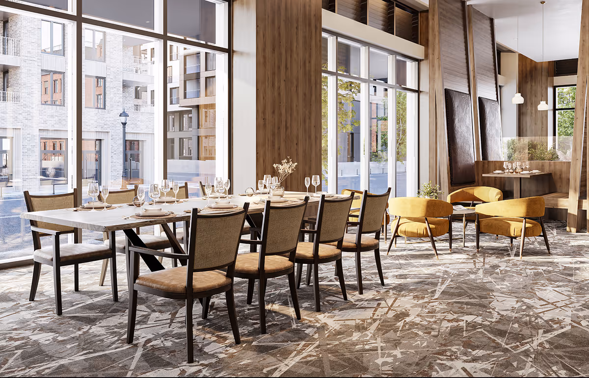 A bright dining room with a long table set with plates, glasses, and cutlery. The room features large floor-to-ceiling windows letting in natural light, modern wooden paneling on the walls, and a patterned carpet. There are several chairs around the table and a seating area with mustard yellow armchairs and a small table in the background.