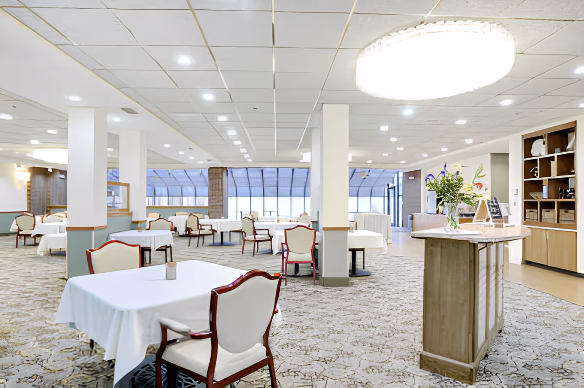 A spacious dining room with multiple tables covered in white tablecloths and surrounded by chairs with wooden frames and white cushions. The room has a patterned carpet, white walls with pillars, and large windows in the background allowing natural light. A round chandelier hangs from the ceiling, and a wooden counter with flowers and decorative items is visible on the right side.