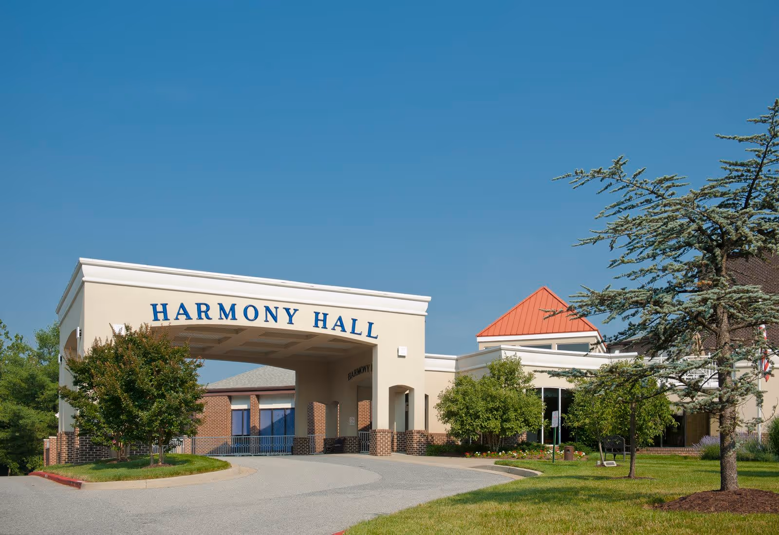 Exterior view of the entrance to Harmony Hall, a senior living facility, featuring a covered driveway with the name 'Harmony Hall' displayed prominently. The building has a beige facade with brick accents, surrounded by green trees and a well-maintained lawn under a clear blue sky.