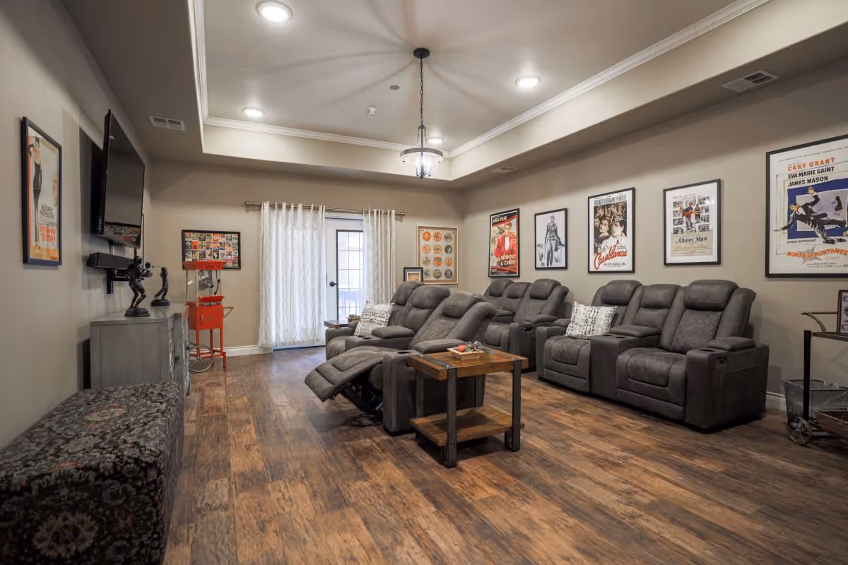 A cozy living room with multiple gray reclining chairs arranged in two rows facing a wall-mounted flat screen TV. The room has wood flooring, a small wooden coffee table, and movie posters framed on the walls. A popcorn machine is visible near a glass door with sheer white curtains.
