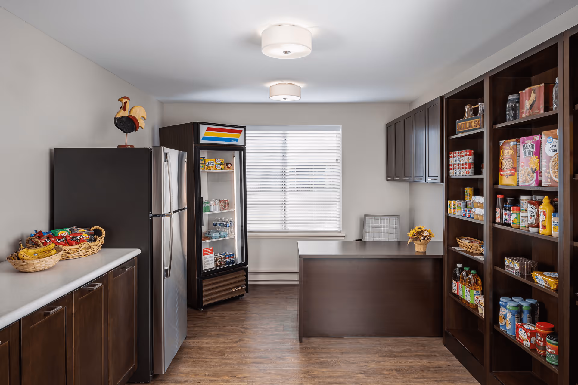 A small pantry or snack room with dark wood cabinets and shelves stocked with various food items and snacks. There is a stainless steel refrigerator, a glass-front beverage cooler, a countertop with baskets of chips and bananas, and a desk with a chair near a window with blinds. The room has wood flooring and two ceiling lights.