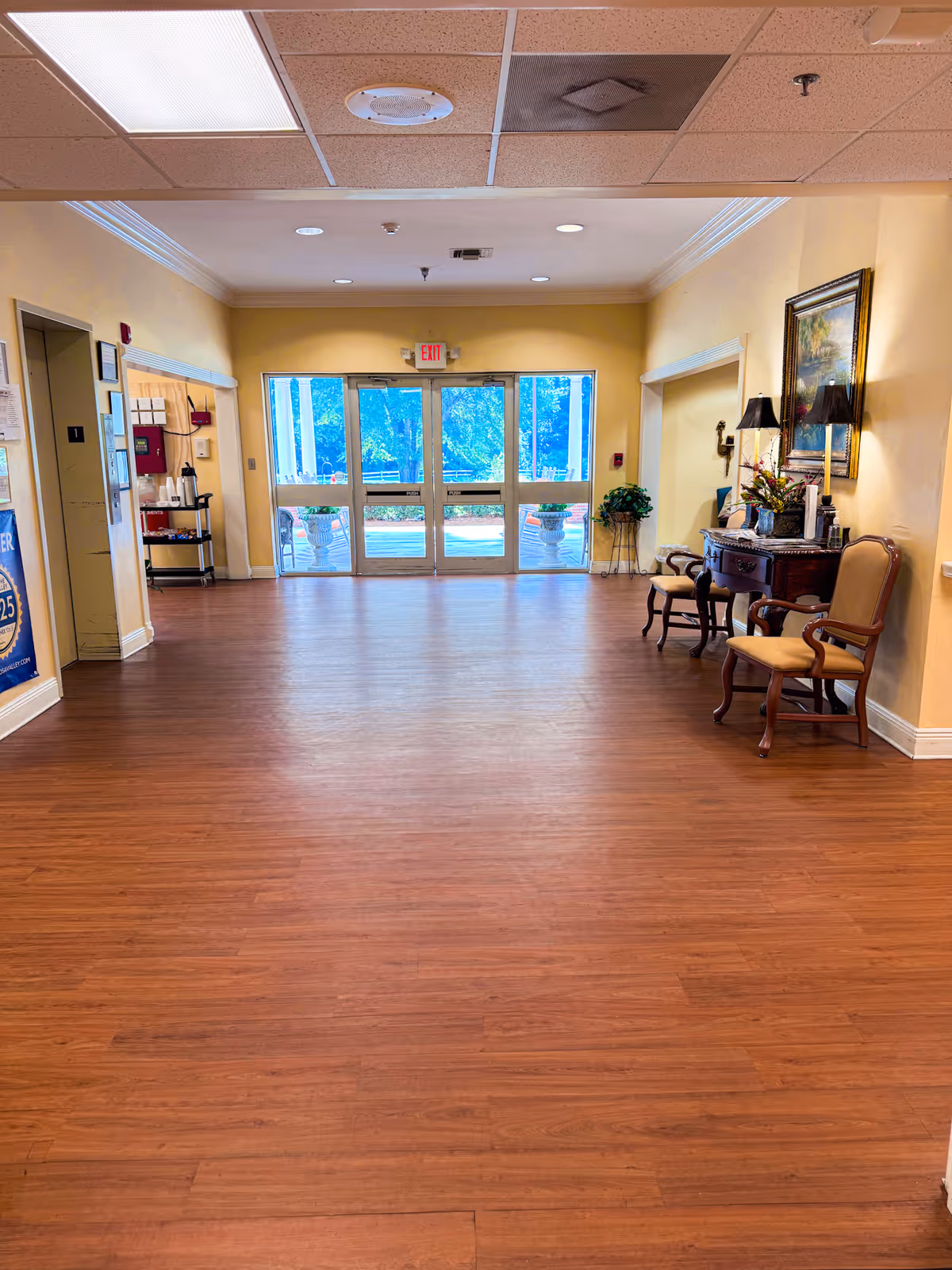 A spacious hallway with wooden flooring and yellow walls in an assisted living facility. There are double glass exit doors at the far end letting in natural light. On the right side, there are two wooden chairs with cushions and a small table with a lamp, flowers, and a framed painting above it. On the left side, there is a small shelf with coffee supplies and a bulletin board on the wall.