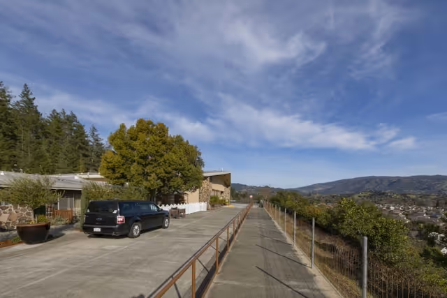 Exterior view of Cloverdale Healthcare Center showing a paved driveway with a parked black vehicle, a large tree near the building entrance, and a scenic view of hills and a residential area under a partly cloudy sky.