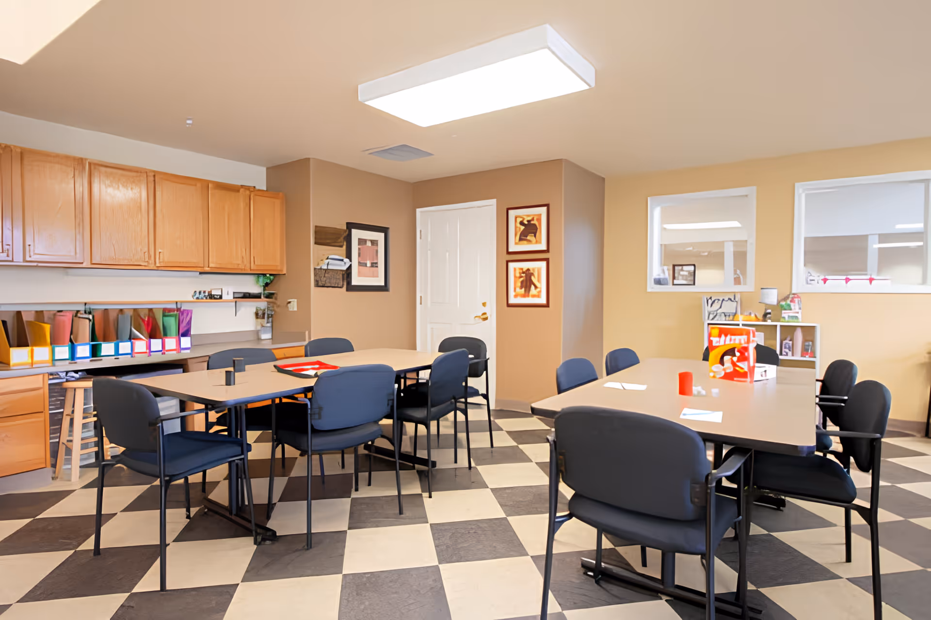 A well-lit room with checkered black and white flooring, featuring two tables surrounded by black chairs. The room has beige walls with framed artwork, wooden cabinets along one wall, and shelves with colorful folders and office supplies. There are two windows on one wall looking into another room.