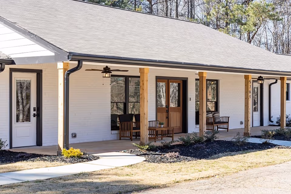 Exterior view of a single-story senior living facility building with a covered porch supported by wooden posts. The porch has ceiling fans and outdoor seating with chairs and a small table. The building has white brick walls, multiple doors and windows, and landscaped areas with shrubs and mulch.