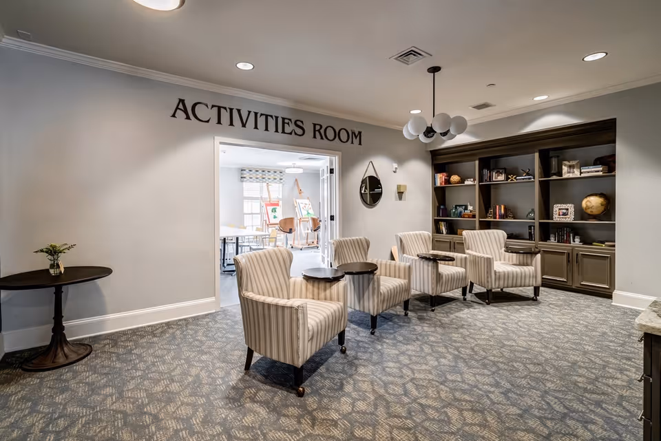 A cozy seating area with four striped armchairs each paired with a small round side table, positioned in front of a built-in bookshelf filled with books and decorative items. The wall above the doorway reads 'ACTIVITIES ROOM'. Through the open double doors, an adjoining room with tables, chairs, and easels with paintings is visible. The room has soft lighting, a patterned carpet, and neutral-colored walls.