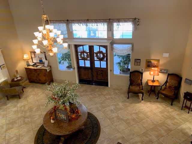 View of a spacious, well-lit lobby area with a large chandelier hanging from the ceiling. There is a round table in the center with a floral arrangement and decorative items. The entrance features double wooden doors with glass panels and wreaths. Two chairs and a small table with a lamp are placed against the wall to the right. A wooden cabinet with a lamp and some papers is on the left side near a small green chair. The floor is tiled with a beige pattern.
