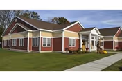 Exterior view of a single-story senior living facility building with red siding and white trim, surrounded by a well-maintained lawn and a concrete walkway leading to the entrance.