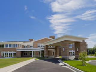 Brick senior living facility with a covered entrance, driveway, and green lawn under a blue sky.