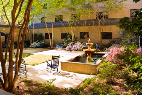 A peaceful outdoor courtyard at Westminster Towers featuring a central water fountain surrounded by a small pond. There are several rocking chairs placed on the paved walkway, with trees, shrubs, and flowering plants providing greenery and shade. The building's brick exterior and windows are visible in the background.