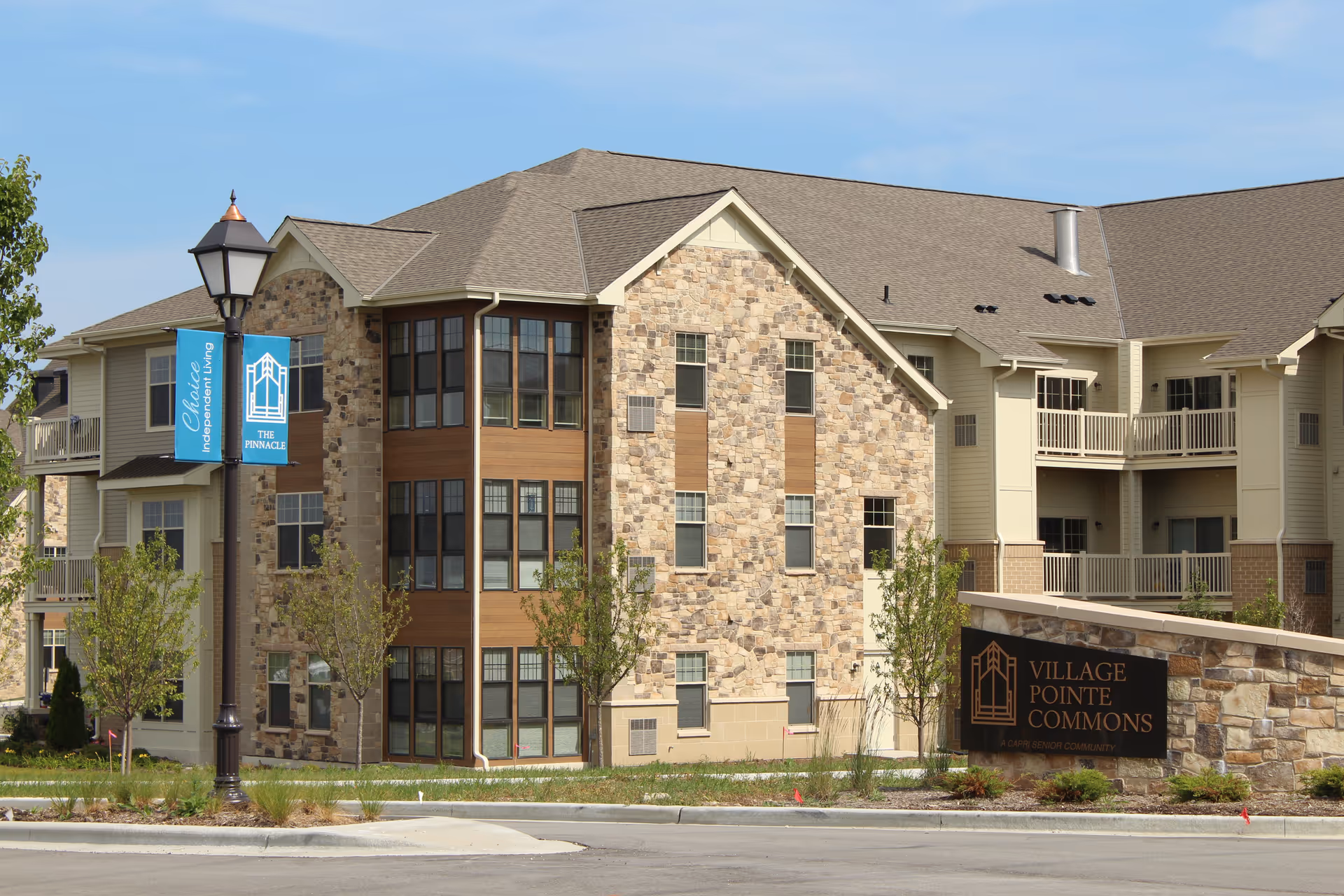 Exterior view of a stone-and-siding senior living building with balconies and a sign reading 'Village Pointe Commons'.
