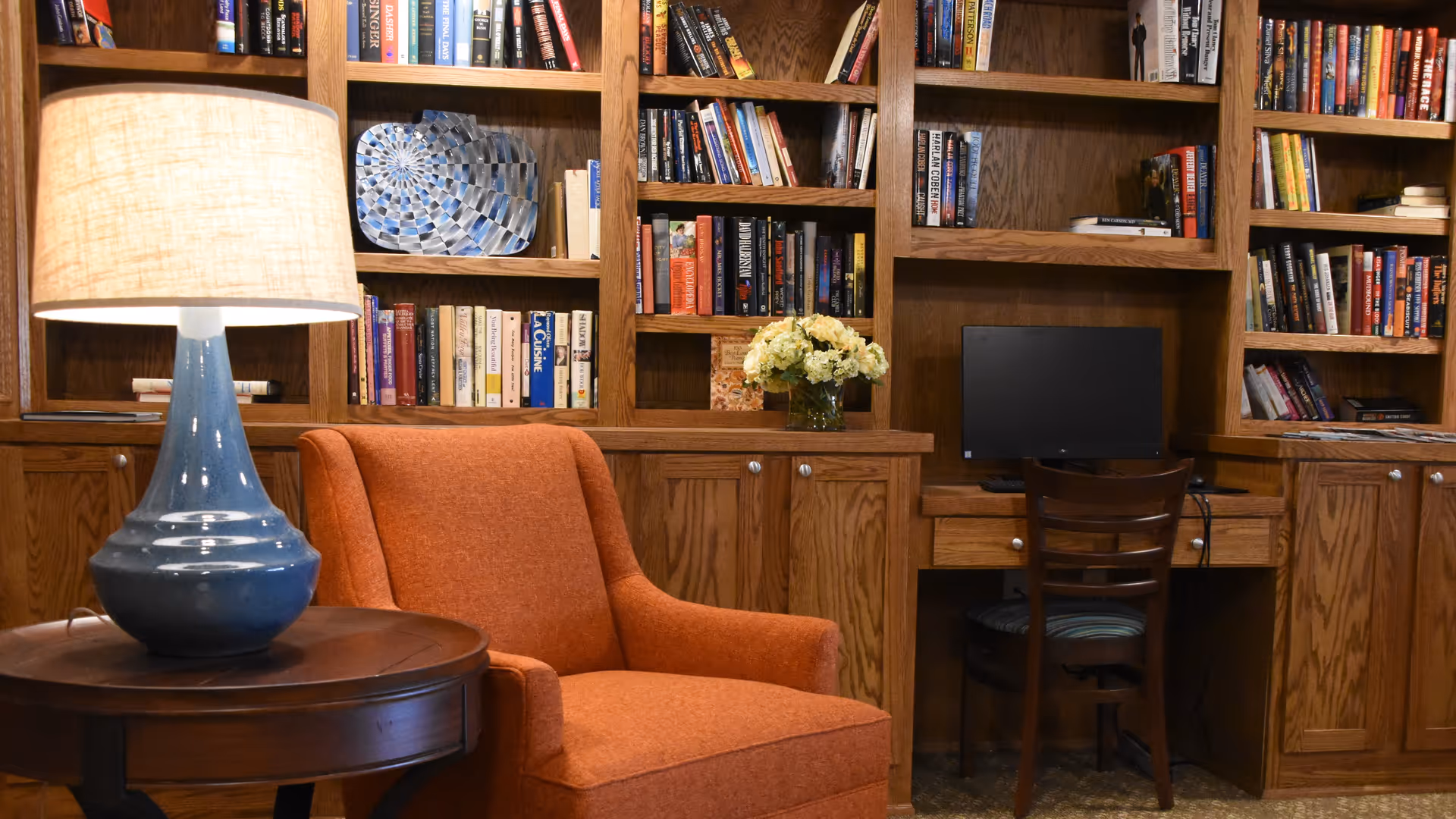 A cozy reading area with an orange armchair, a side table and lamp in front of wooden bookshelves and a small desk with a computer.