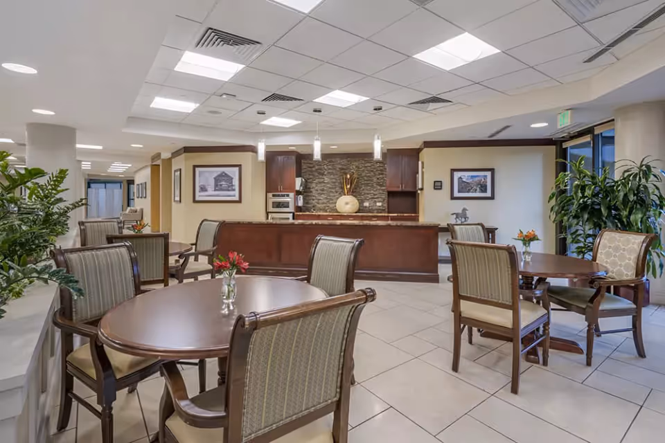 A well-lit dining area in a senior living facility with round wooden tables and cushioned chairs. The room features a kitchen counter with pendant lights, framed pictures on the walls, and several green plants adding a touch of nature. The floor is tiled, and the ceiling has recessed lighting and ventilation panels.