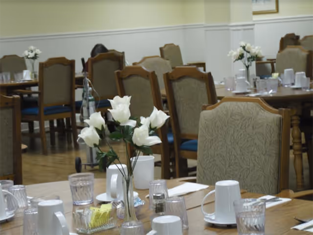 Communal dining room with multiple tables and chairs set with white mugs, glasses, and vases of white flowers.