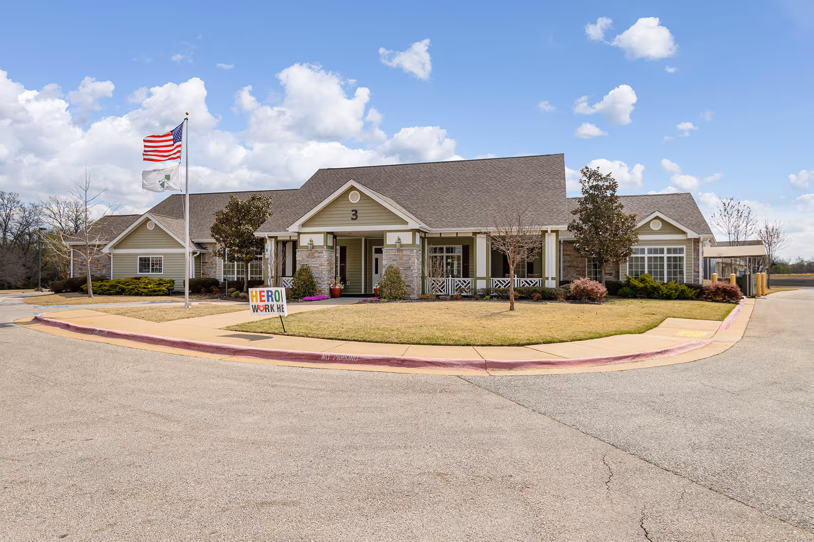Exterior view of a single-story assisted living facility building with a peaked roof, beige siding, and stone accents. There are two flagpoles in front, one with an American flag and the other with a white flag. A small lawn with a sidewalk surrounds the building entrance, and a colorful sign that reads 'HERO WORK HE' is placed near the sidewalk. The sky is partly cloudy.
