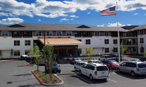 Exterior view of a multi-story senior living facility with a covered entrance, several parked cars in the parking lot, small trees planted along the driveway, and an American flag flying on a flagpole against a partly cloudy sky.