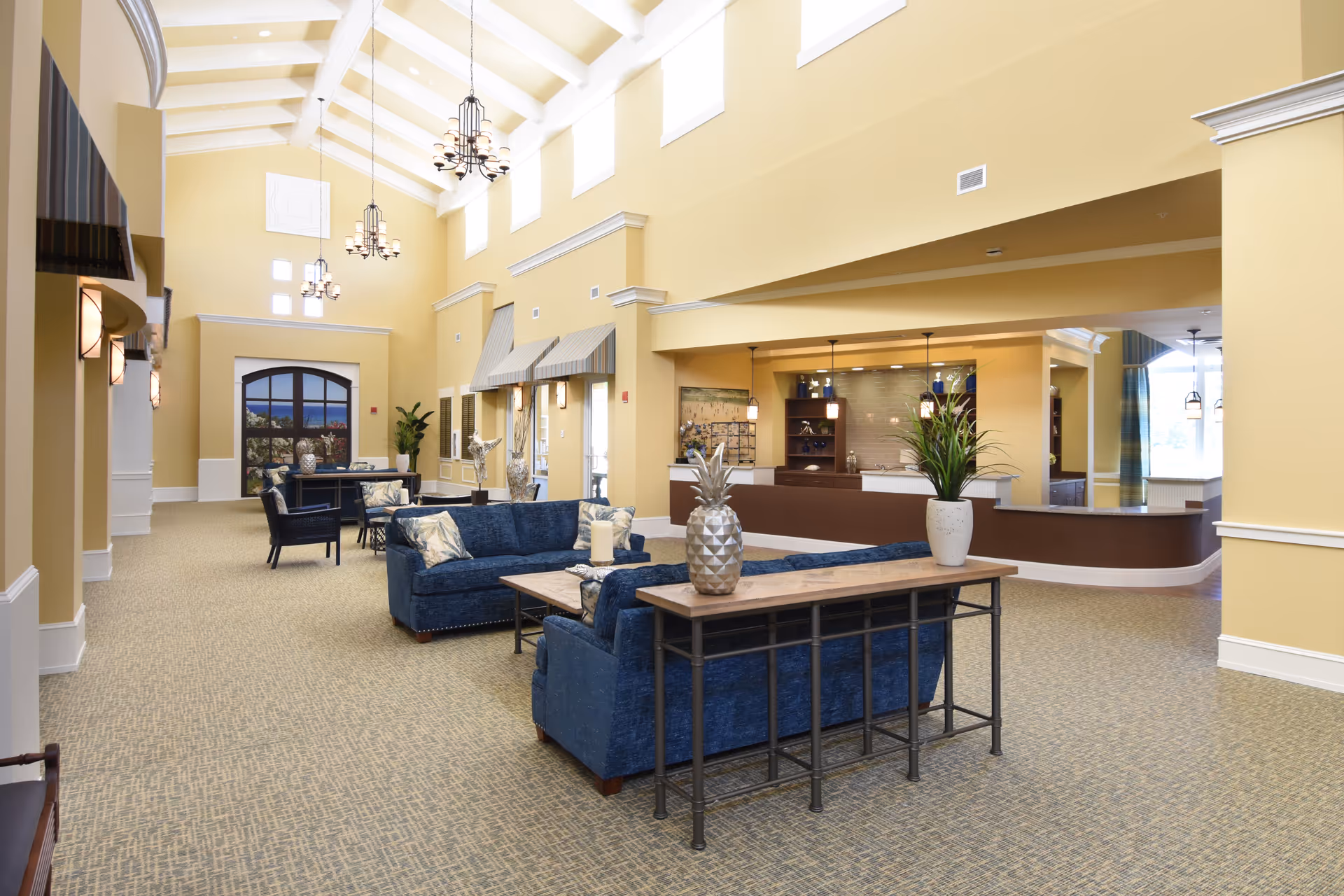 Spacious senior living facility common area with high vaulted ceilings, yellow walls, and multiple hanging chandeliers. The room features blue upholstered sofas with patterned cushions, a wooden console table with decorative plants and a pineapple ornament, and a reception desk area in the background. Large windows near the ceiling allow natural light to fill the space.
