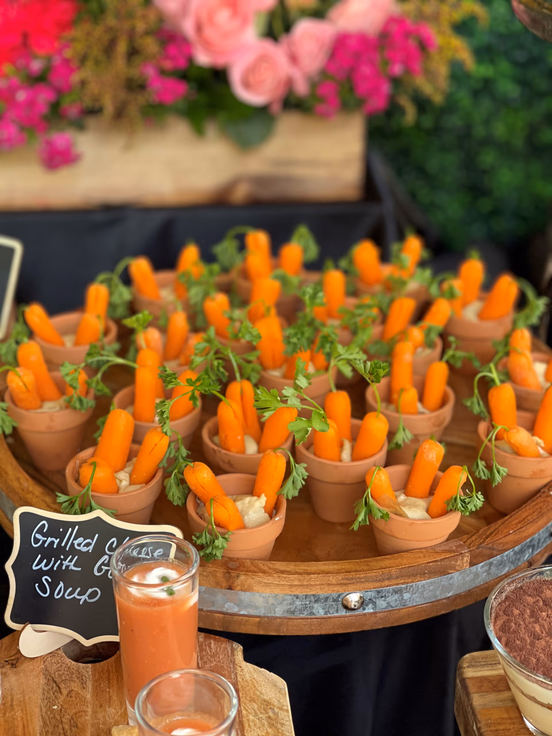 Small terracotta pots filled with dip and baby carrots arranged on a wooden tray, with a small chalkboard sign that reads 'Grilled Cheese with Soup' and two glasses of tomato soup in the foreground. Pink and red flowers are blurred in the background.