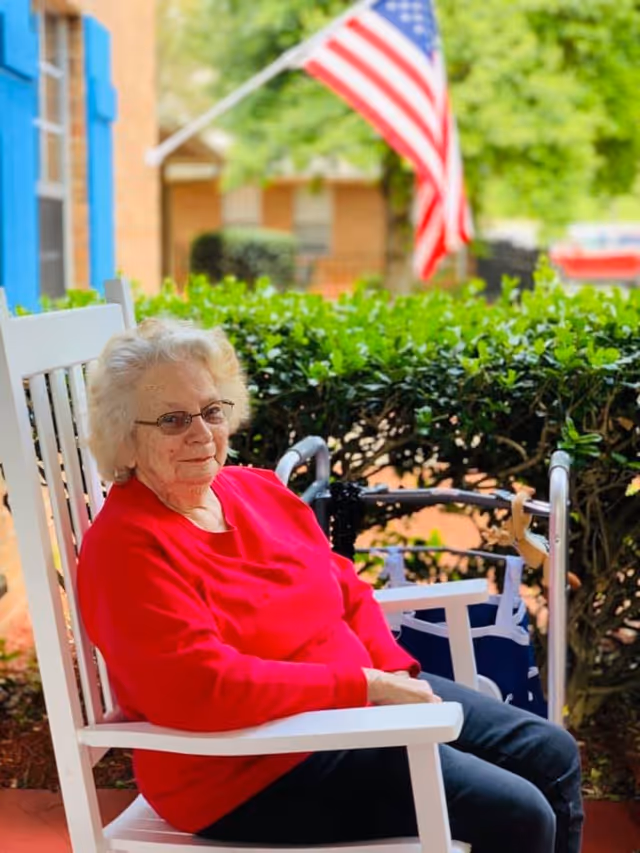 An elderly woman wearing glasses and a red sweater sits on a white rocking chair on a porch. Behind her, there is a green hedge, an American flag, and a walker. The background includes a brick building with blue window shutters.