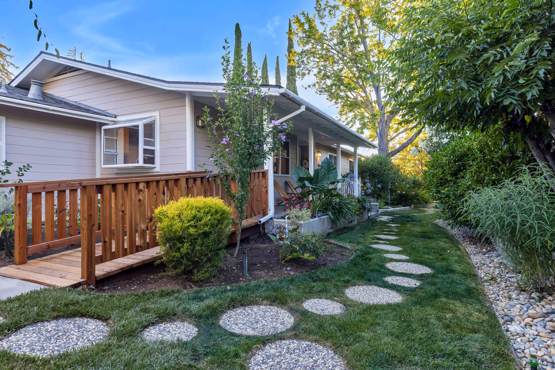 A well-maintained garden path with circular stone stepping stones leading alongside a single-story building with a wooden ramp and porch. The garden features various green shrubs, flowering plants, and tall trees under a clear blue sky.