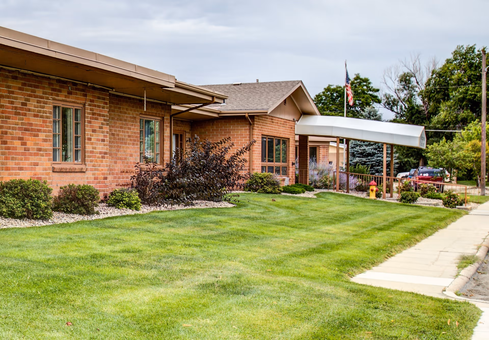 Brick single-story building with a covered entrance canopy, manicured lawn, shrubs, and an American flag.