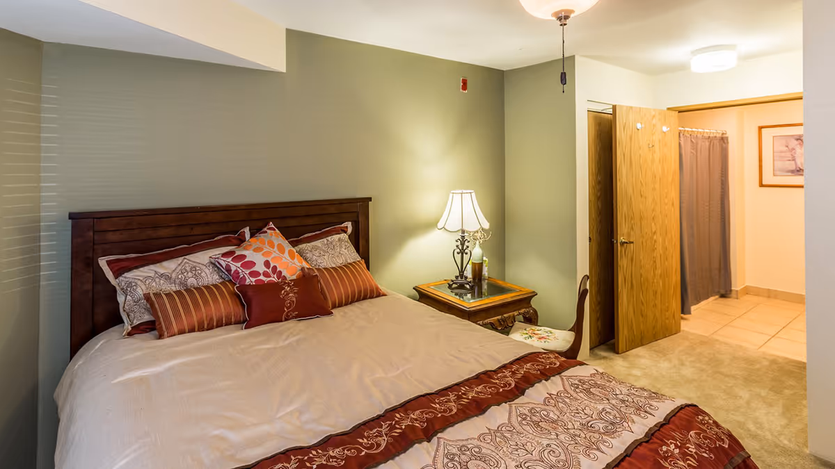 Cozy bedroom with a large bed topped with decorative pillows, a wooden nightstand and lamp, and an open doorway leading to a tiled area.