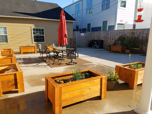 Sunny outdoor courtyard with wooden planter boxes, a patio table and chairs under a red umbrella, and surrounding buildings.