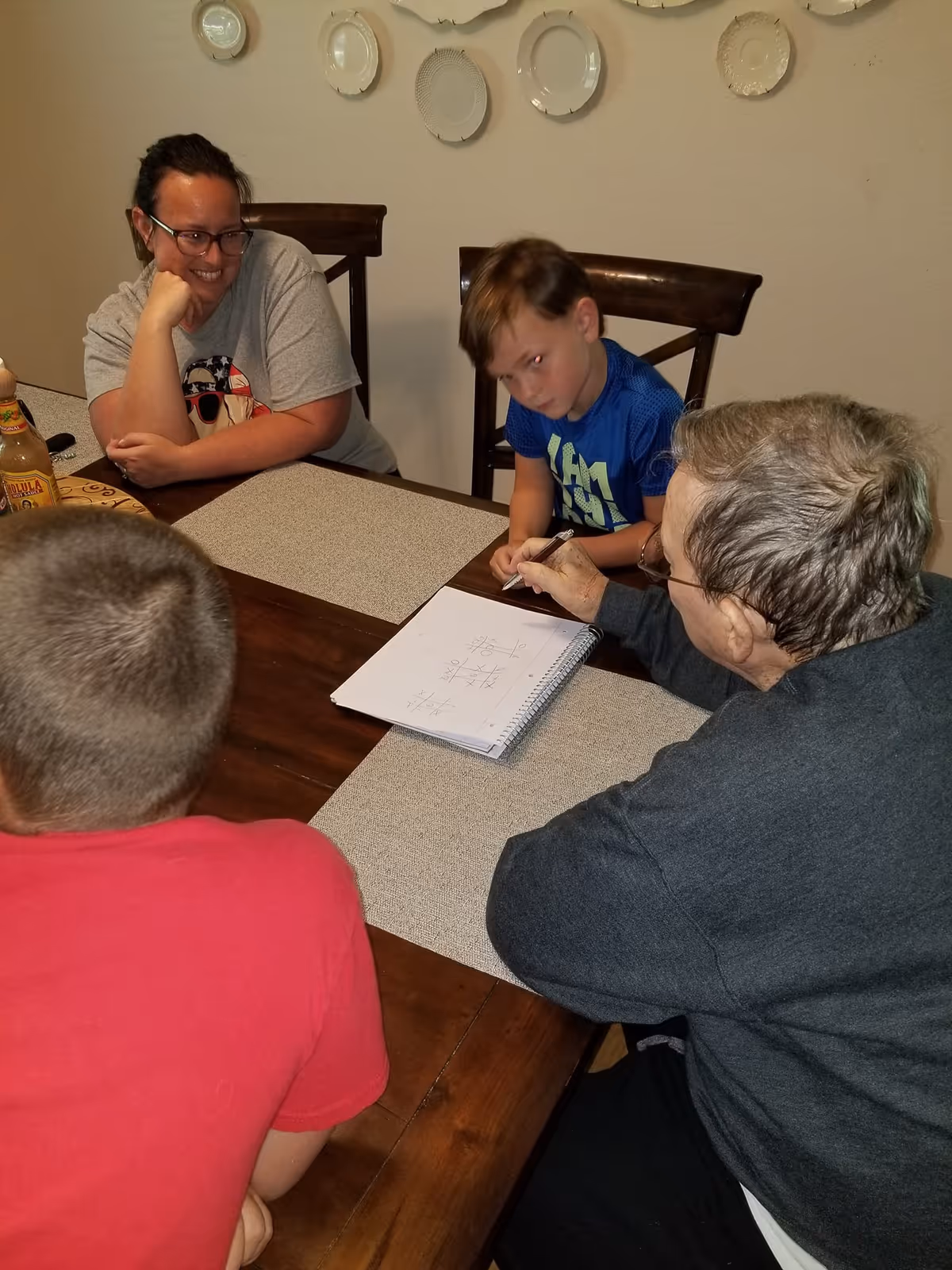 An elderly person and three children sitting around a wooden dining table playing tic-tac-toe on a notepad. The elderly person is holding a pen and appears to be making a move. A woman is smiling and watching the game. Decorative plates are hung on the wall in the background.