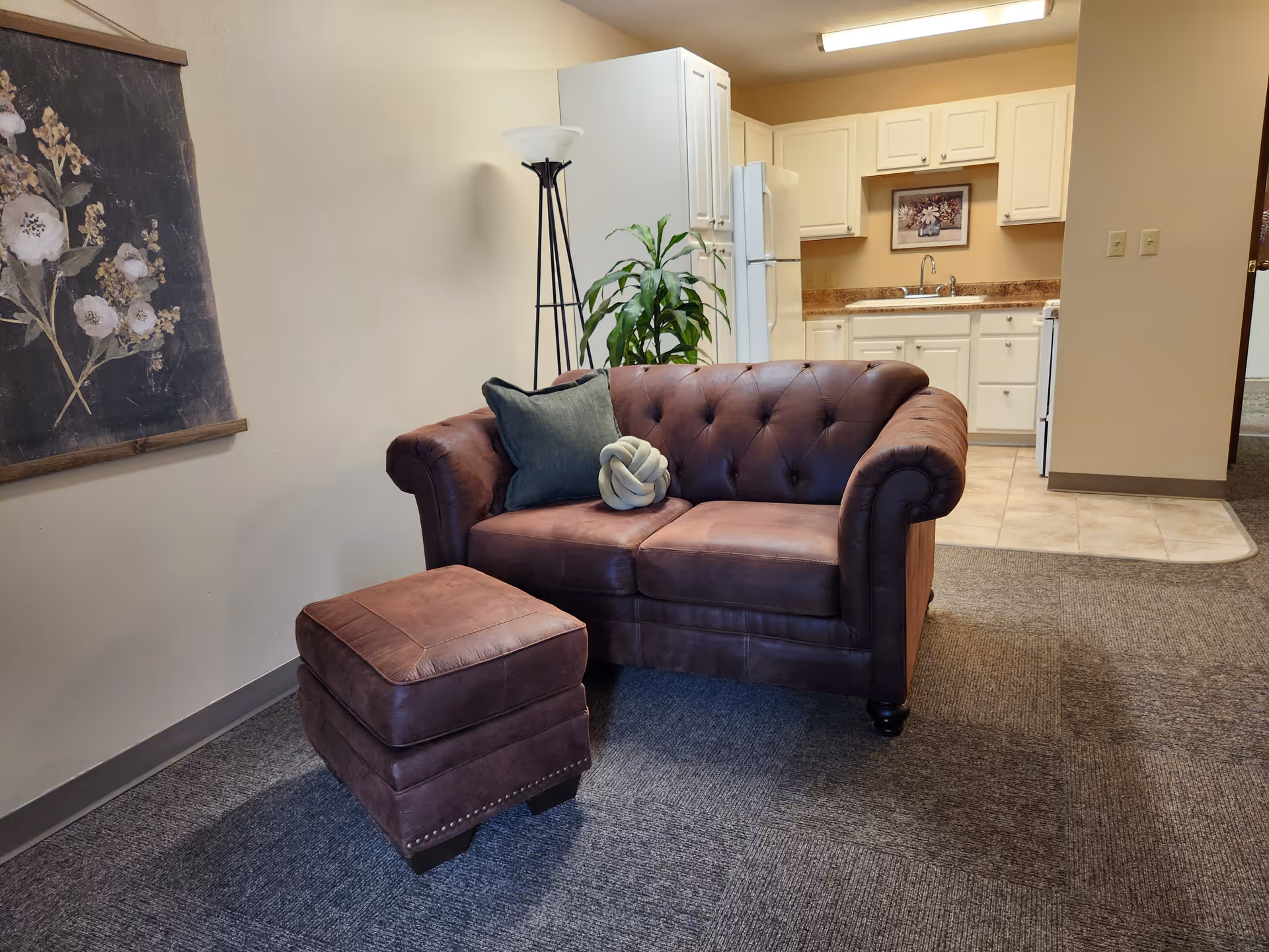 A cozy living area with a brown leather tufted loveseat and matching ottoman on a carpeted floor. There are two decorative pillows on the loveseat, one green and one knotted gray. Behind the loveseat is a tall floor lamp and a green potted plant. In the background, there is a small kitchen area with white cabinets, a refrigerator, a sink, and a stove. A floral painting hangs on the wall to the left.
