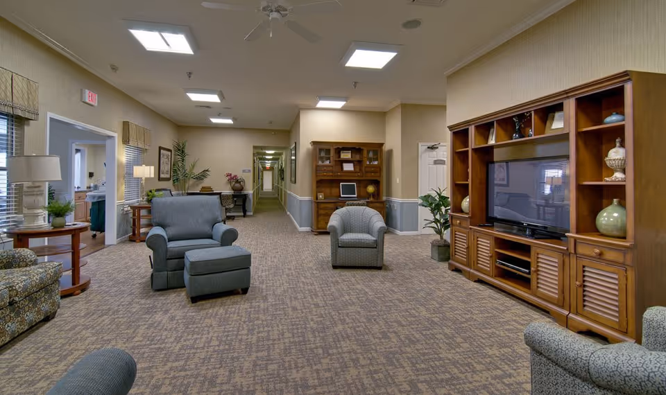 A spacious senior living common area with patterned carpet, several upholstered armchairs, a large wooden entertainment center with a flat-screen TV, and a wooden desk with a computer. The room is well-lit with ceiling lights and has beige walls with blue wainscoting. There is a hallway leading further into the facility and windows with blinds on the left side.