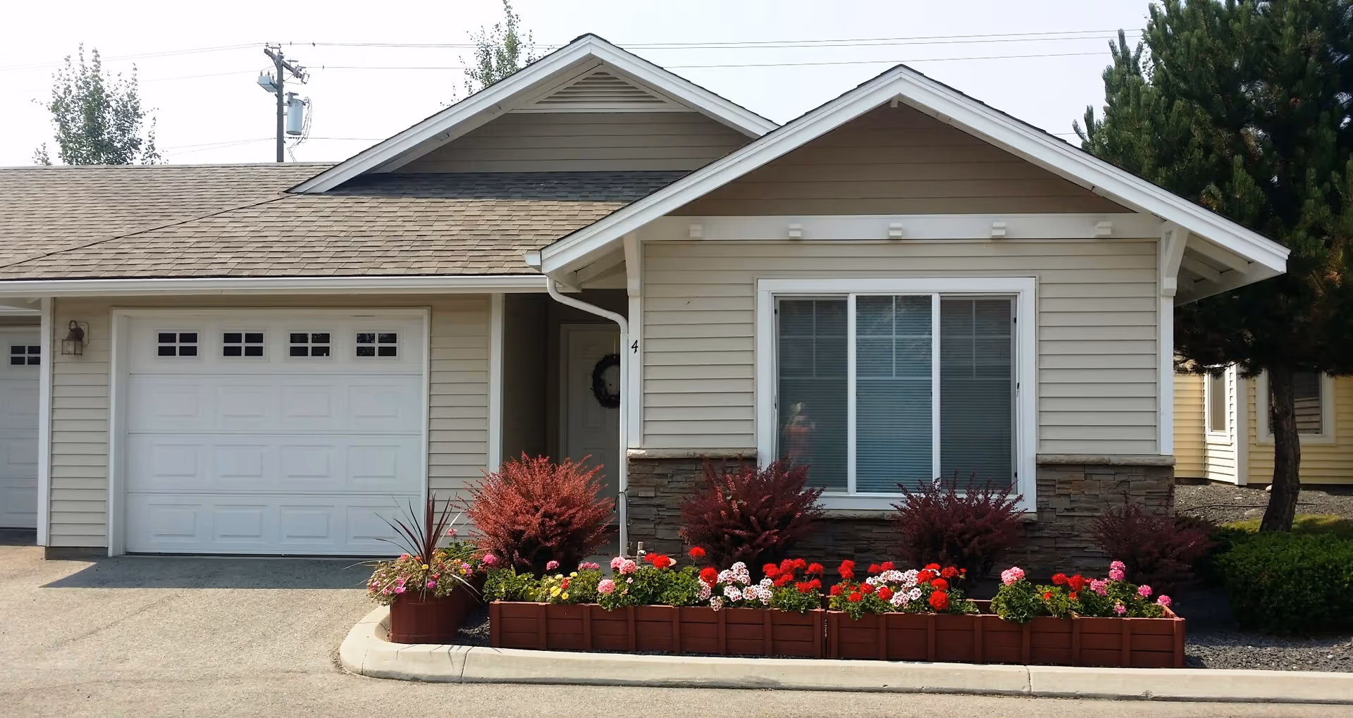 Exterior view of a single-story independent living cottage with beige siding, a white garage door, a large window with blinds, and a small garden bed filled with colorful flowers and shrubs in front.