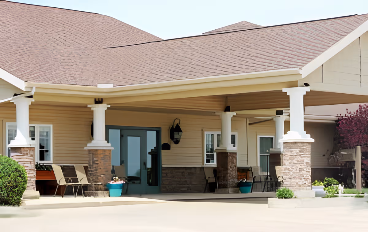 Entrance area of a senior living facility with a covered drop-off zone supported by white columns with stone bases. There are chairs and potted plants near the entrance doors, and the building has beige siding with a brown shingled roof.