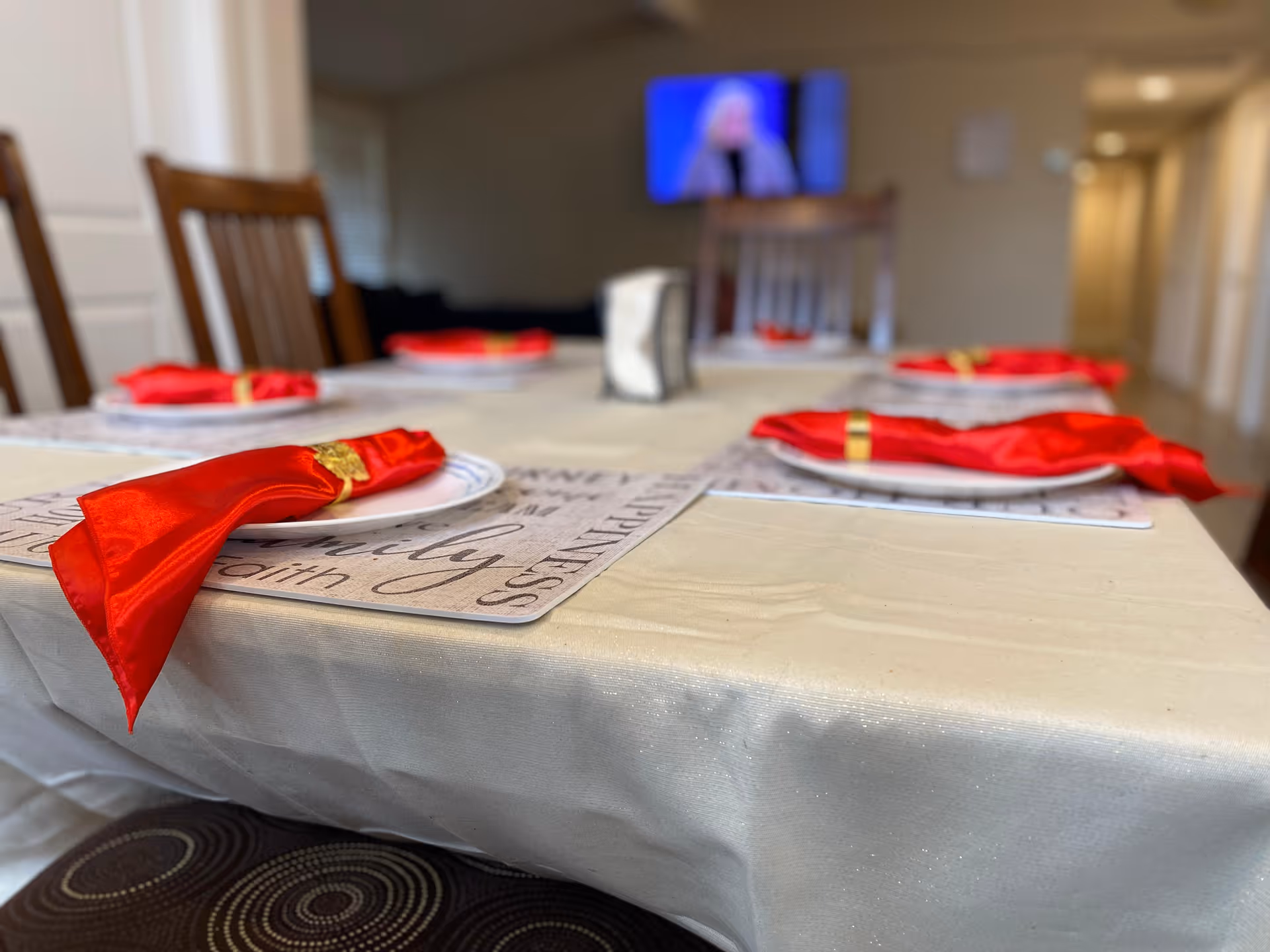 Dining table set with plates and red napkins and gold rings in a dining area with chairs and a TV in the background.