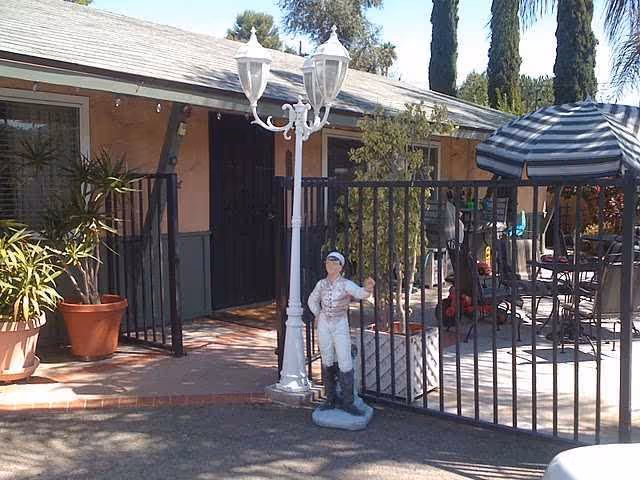 Outdoor patio area of a facility with a black metal fence and gate, a white decorative street lamp, a small statue of a person in riding attire, potted plants, and patio furniture including a table with a striped umbrella.