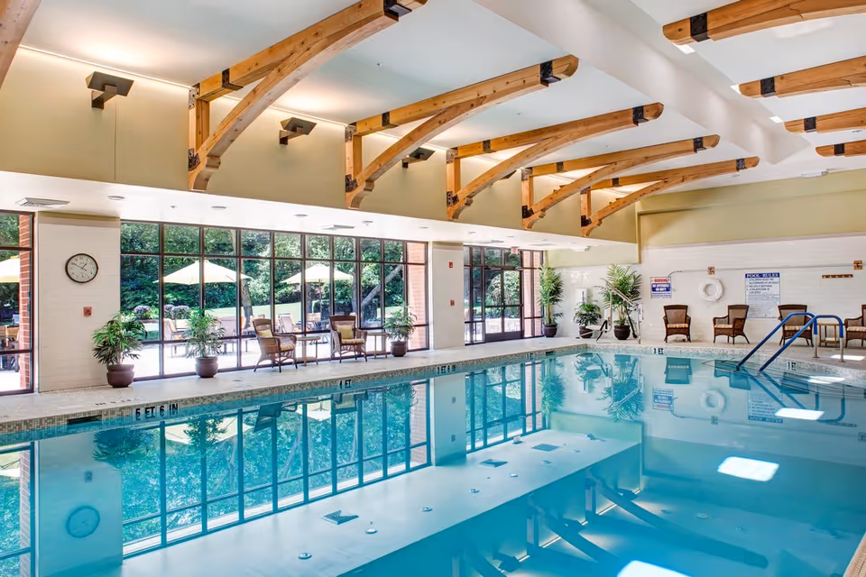 Indoor swimming pool room with exposed wooden ceiling beams, lounge seating, large windows and potted plants.