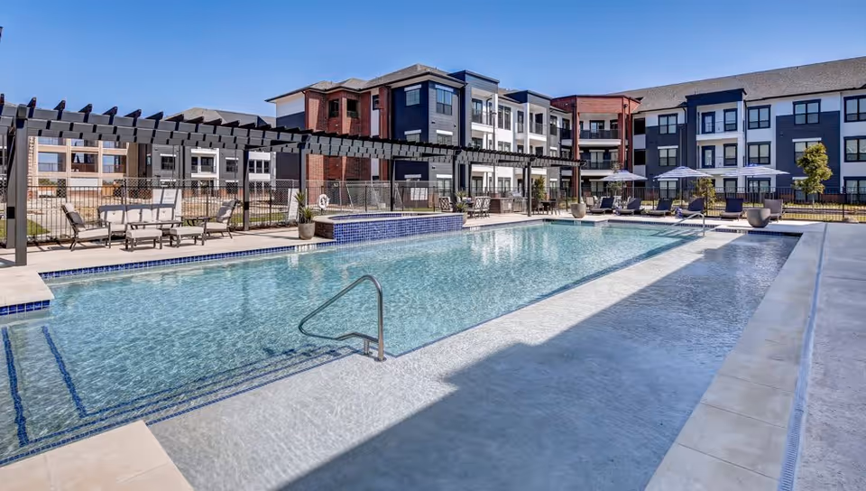 Outdoor swimming pool with clear water surrounded by a modern apartment complex. There are lounge chairs, tables with umbrellas, and a pergola with seating on the pool deck. The sky is clear and blue.
