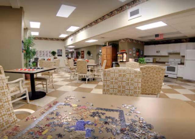 A spacious common area with multiple tables and chairs arranged for seating. The room features a checkered tile floor and a kitchen area with white appliances including a refrigerator, stove, and microwave. Puzzle pieces are spread out on a table in the foreground. The walls are decorated with framed pictures and plants, and the ceiling has recessed lighting.