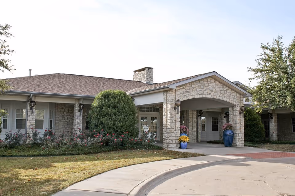 Exterior view of a single-story stone building with a covered entrance, surrounded by greenery and flower pots with colorful flowers.