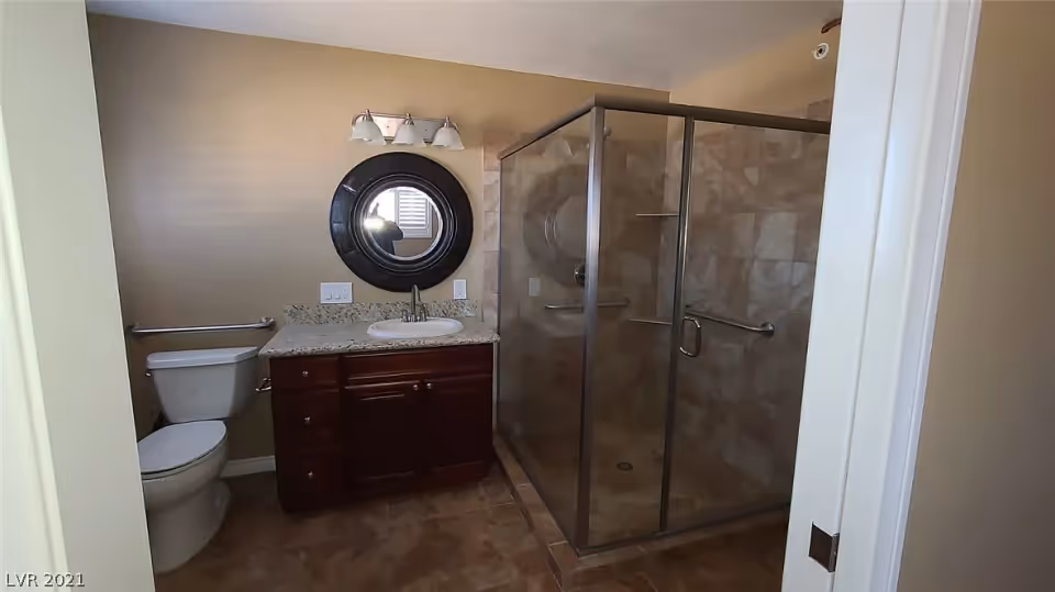 A bathroom with a glass-enclosed shower on the right, a dark wood vanity with a granite countertop and a white sink in the center, a round mirror with a dark frame above the sink, and a toilet on the left side. The walls are beige and the floor is tiled.