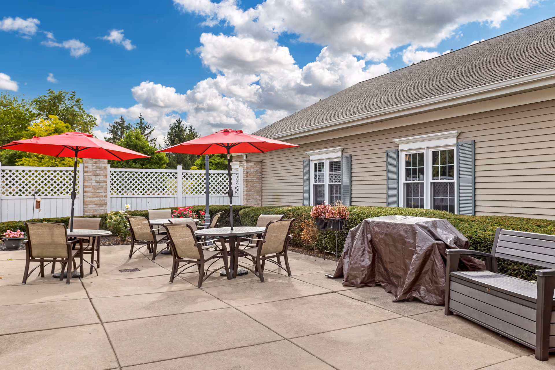 Outdoor patio area with several round tables and chairs, each table shaded by a red umbrella. There is a covered grill and a bench along the side of a beige building with blue shutters on the windows. The patio is surrounded by a white lattice fence and greenery under a partly cloudy blue sky.