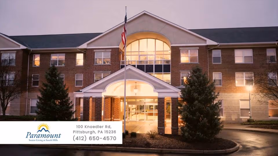 Exterior view of a three-story brick senior living facility building at dusk with lights on inside. The entrance has a covered portico supported by columns, and an American flag is flying on a flagpole in front. There are trees and landscaping around the entrance.