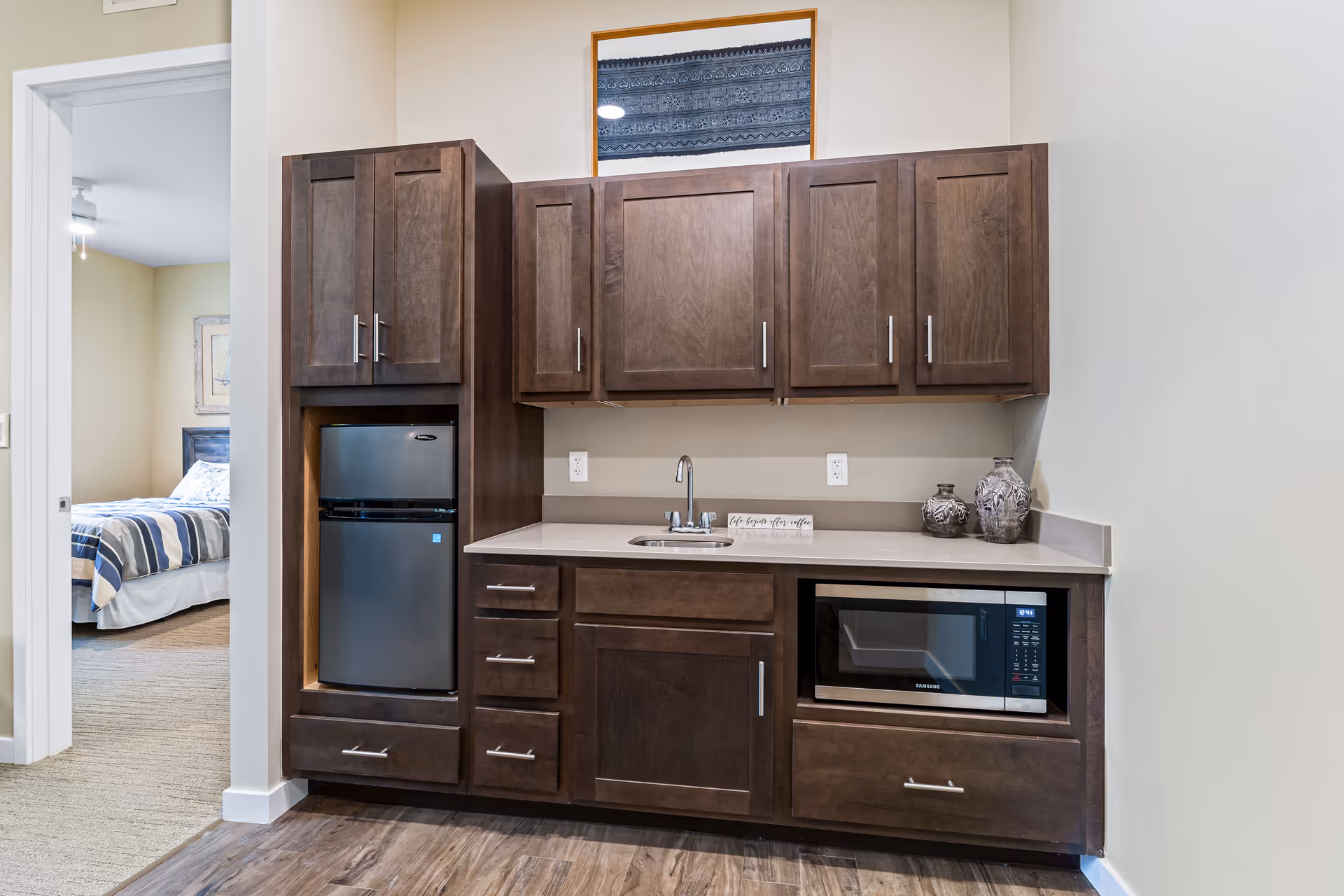 A kitchenette area with dark wood cabinets, a small stainless steel refrigerator, a microwave, a sink, and decorative vases on the countertop. A framed picture hangs above the cabinets. To the left, there is an open doorway leading to a bedroom with a bed covered in striped bedding.