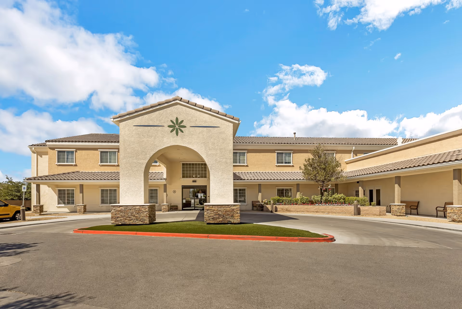 Front entrance of a two-story beige senior living facility with a covered porte-cochere and landscaped driveway under a blue sky.