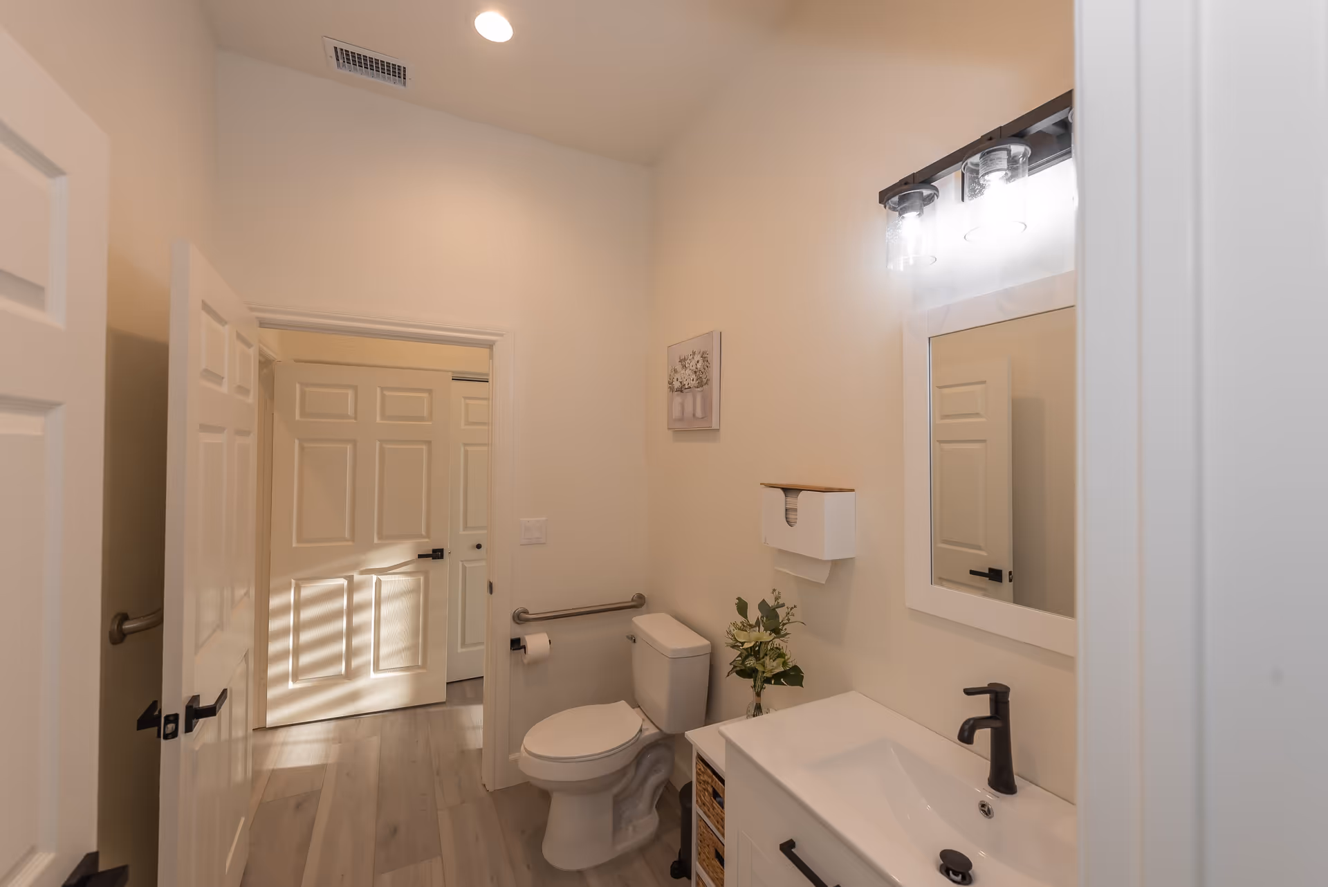 A clean and modern bathroom with light-colored walls and flooring. The bathroom features a white toilet with a grab bar and toilet paper holder on the wall beside it. There is a white vanity with a black faucet and a rectangular mirror above it, illuminated by a two-light fixture. A small vase with flowers is placed on the vanity. The door to the bathroom is open, showing another door in the adjacent room.