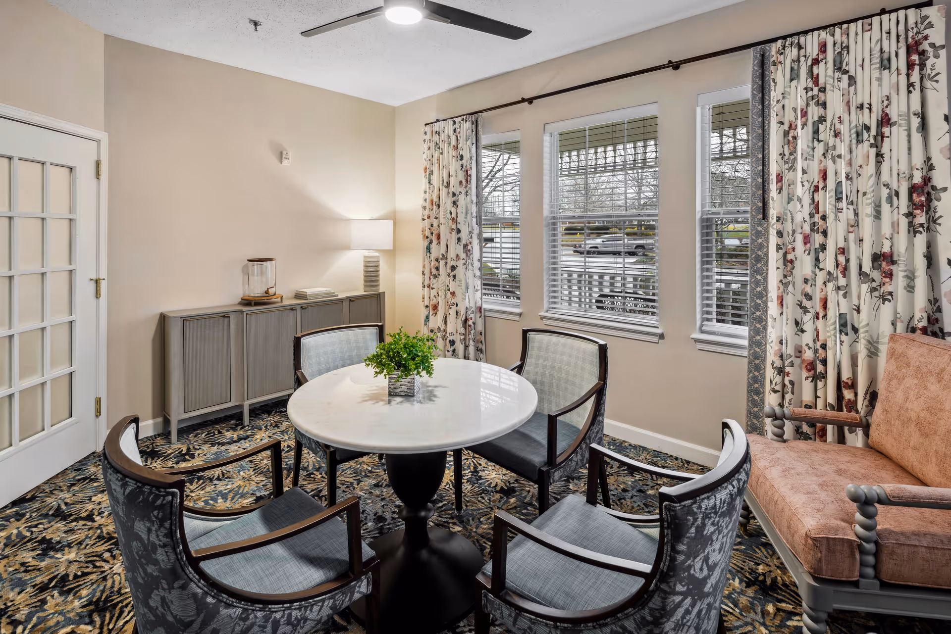 A cozy interior room with a round white table surrounded by four upholstered chairs with wooden arms. There is a small green plant centerpiece on the table. To the right, there is a cushioned bench with wooden armrests. The room has three large windows with white blinds and floral curtains. A sideboard with a lamp and decorative items is against the left wall, and a ceiling fan is visible above.