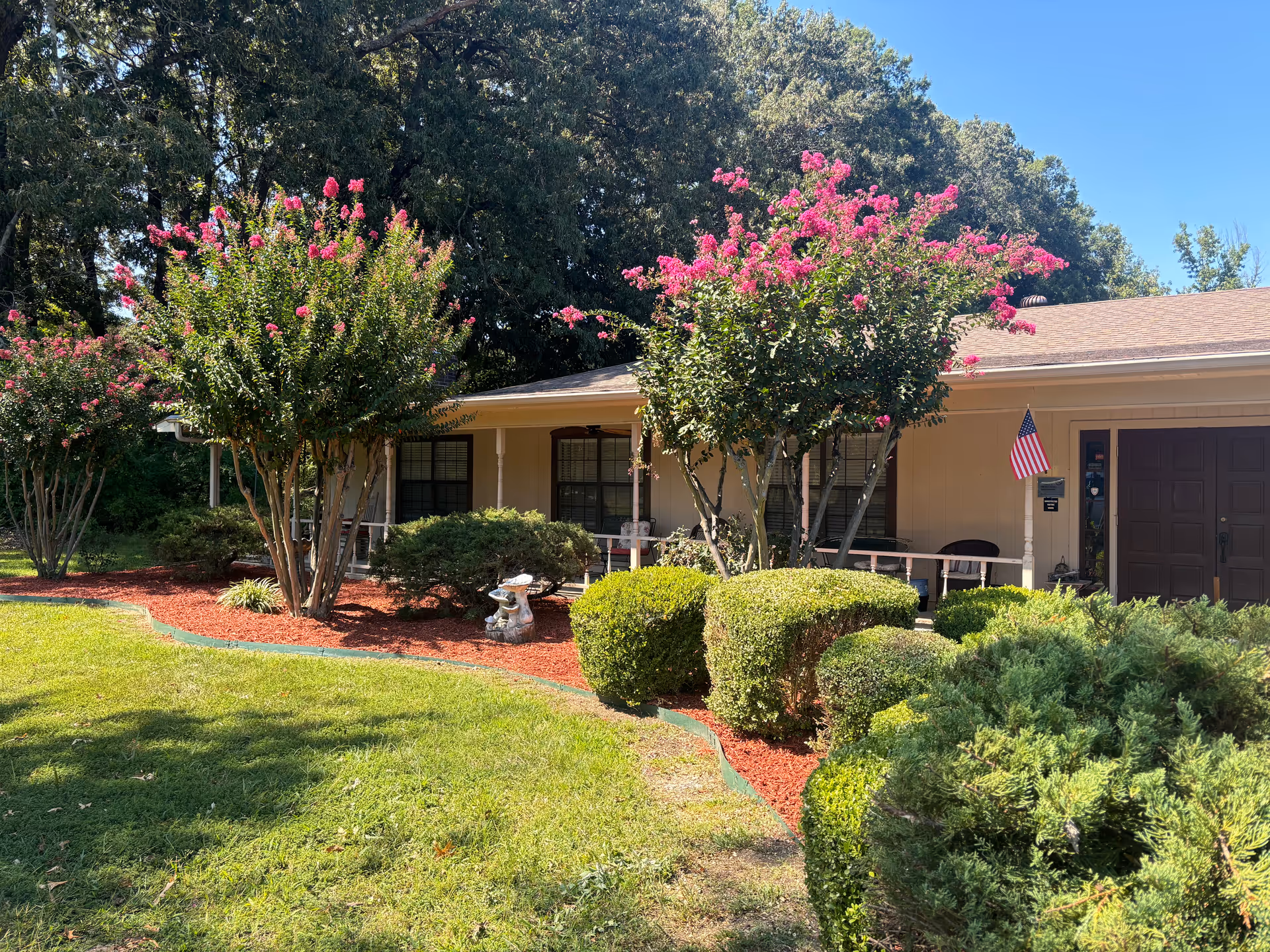 Front exterior view of a single-story building with a porch, surrounded by neatly trimmed bushes and trees with pink flowers, under a clear blue sky.